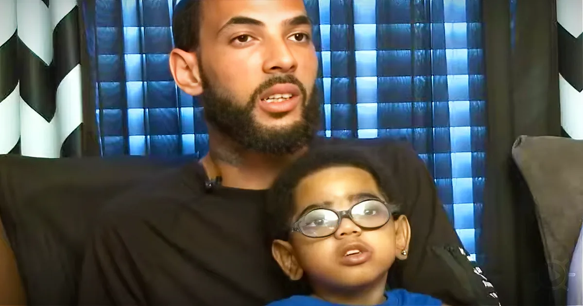 A man with a beard sits behind a child wearing glasses, both on a black couch with a blue checkered background.