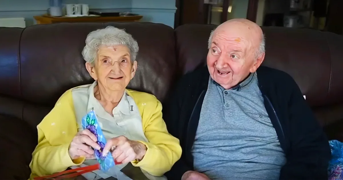 An elderly couple sits on a couch, the woman holding a colorful object, both appearing engaged and cheerful.