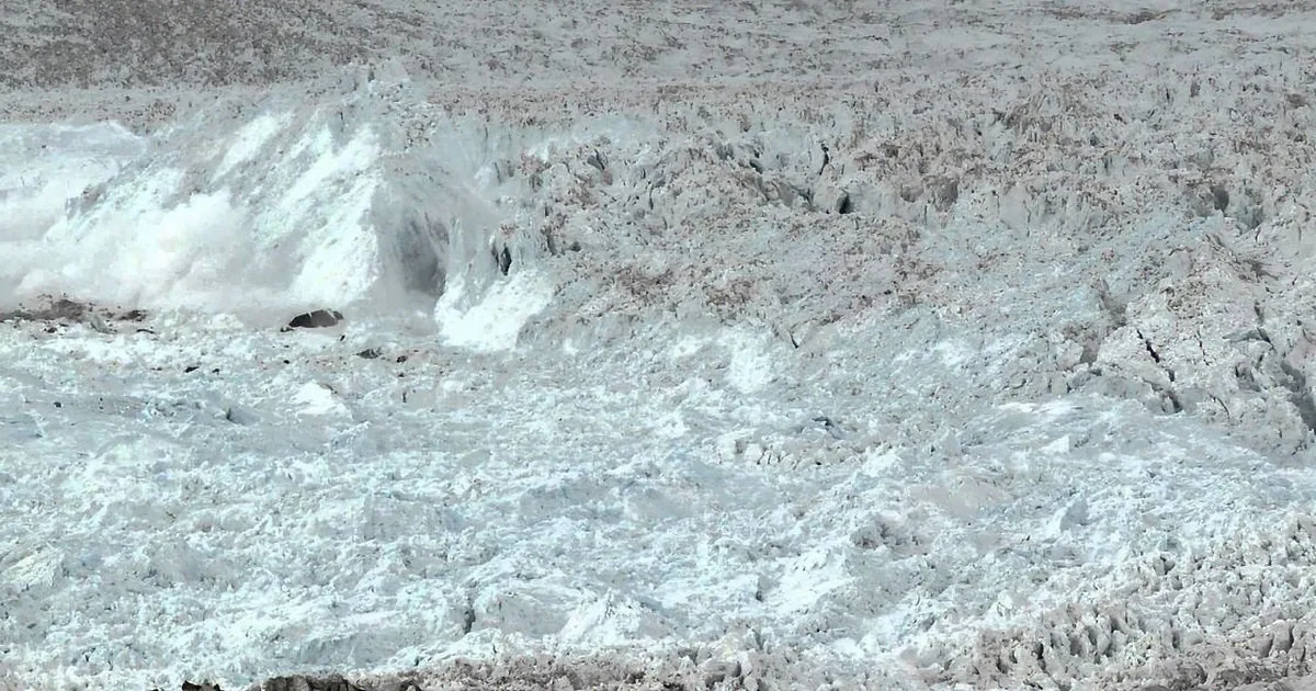 A rugged, icy landscape with a large, textured glacier in the foreground and a snow-covered mountain in the background.