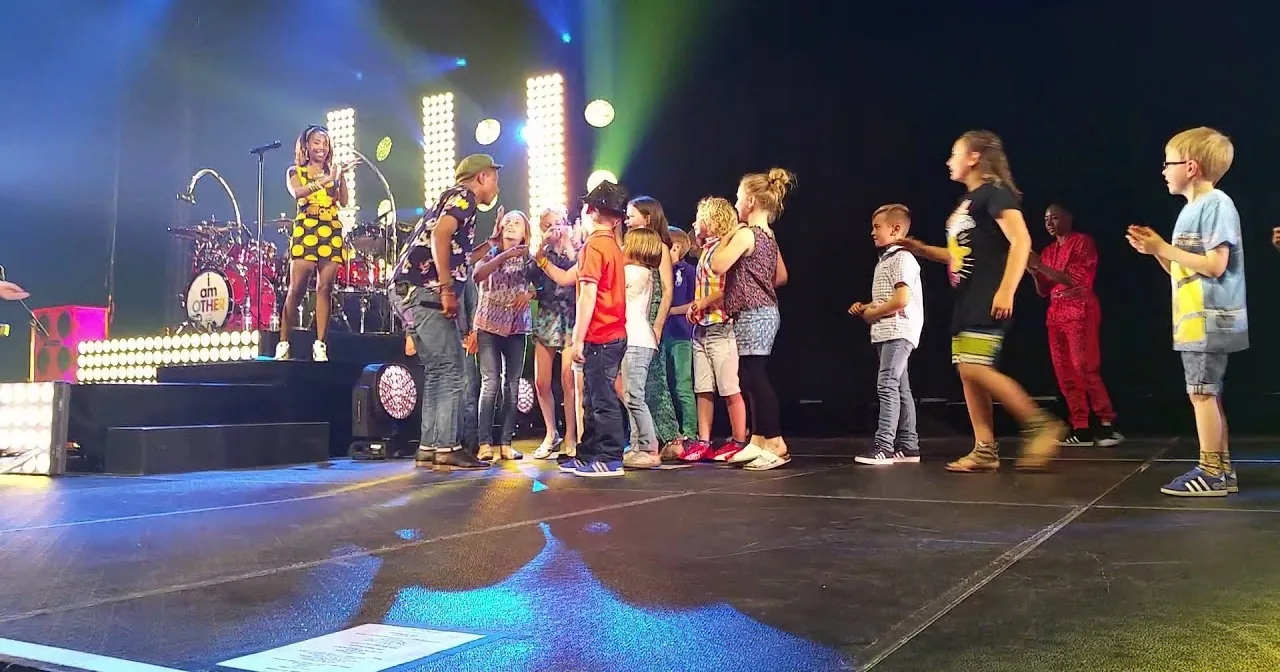 A young woman in a polka dot dress stands on stage, surrounded by children, with colorful stage lights and a drum set in...