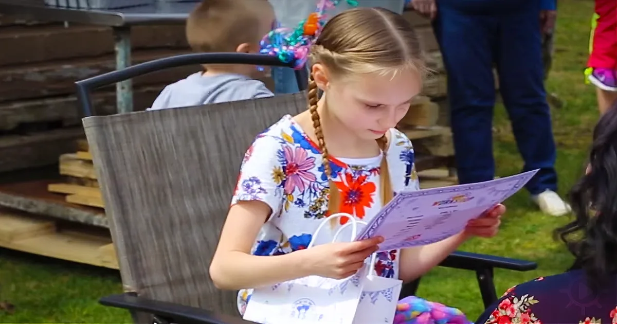 A girl with braids in a floral dress reads a menu while sitting on a chair outdoors.