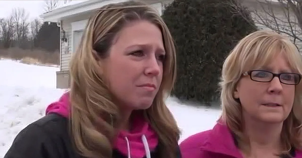 A woman with long blonde hair wearing a pink jacket stands outside in the snow, looking to the side.