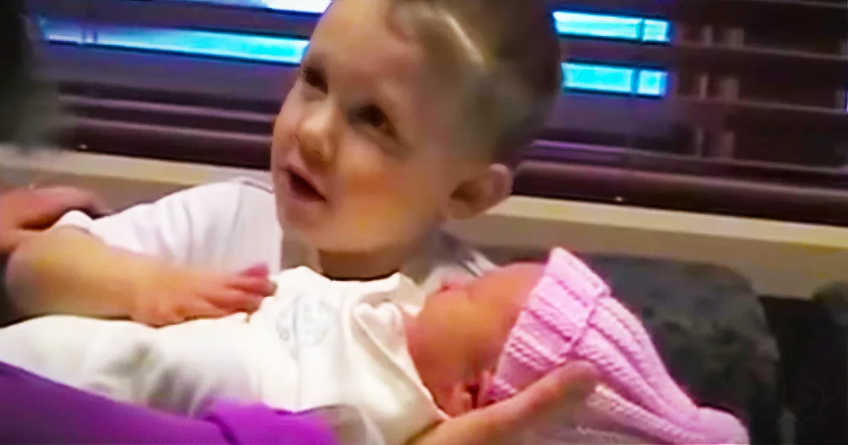 A young child holds a newborn baby in a hospital setting, both wearing white and pink.