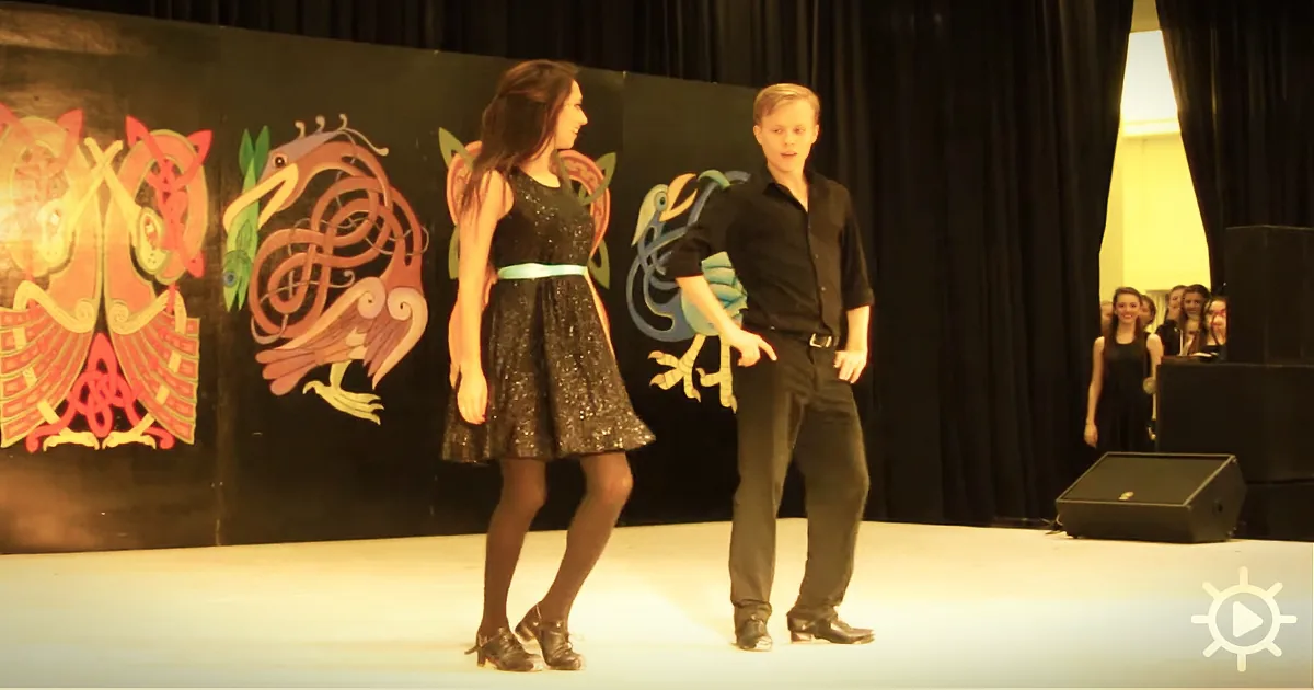 A girl in a black dress and a boy in black stand on stage, facing each other, with colorful bird designs behind them.