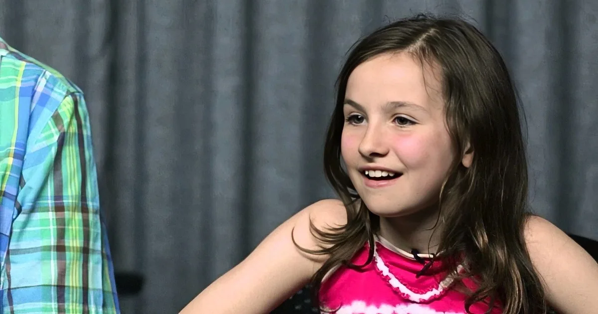 A young girl with long brown hair smiles while wearing a pink and white striped top.