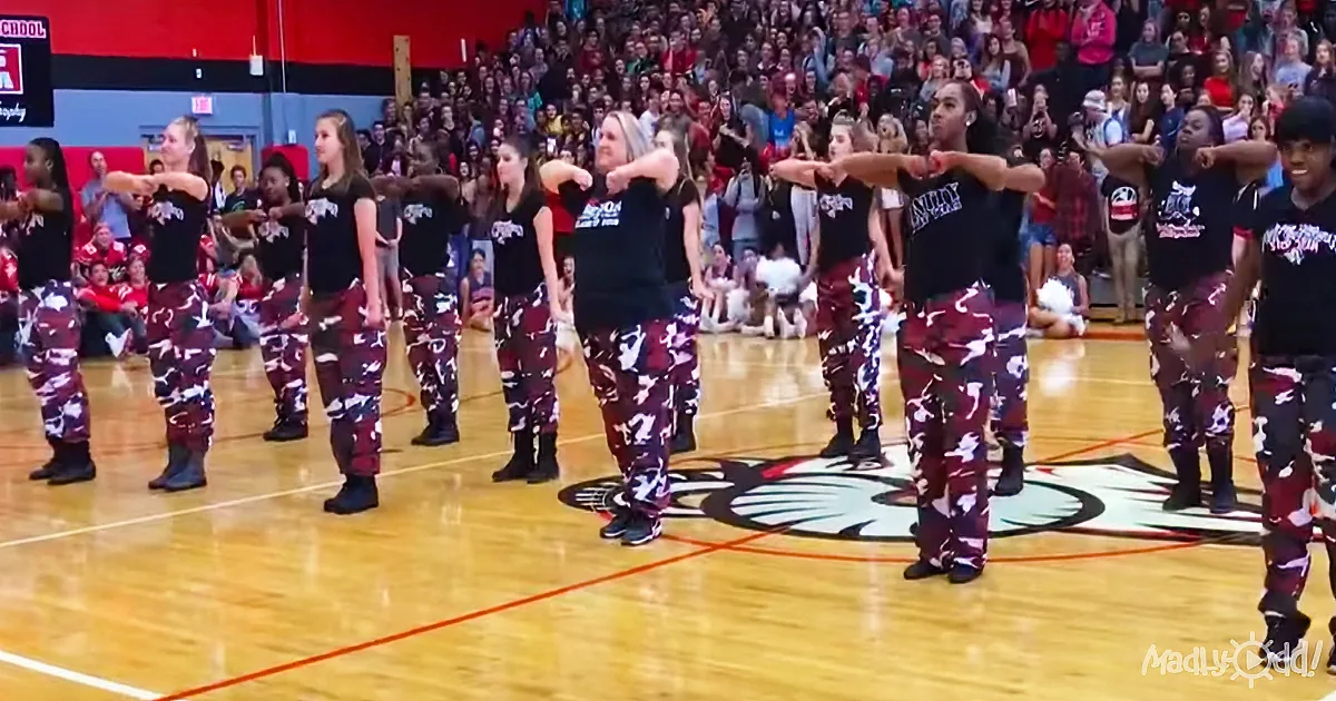 A group of dancers in camouflage pants and black shirts perform on a basketball court in front of a large audience.