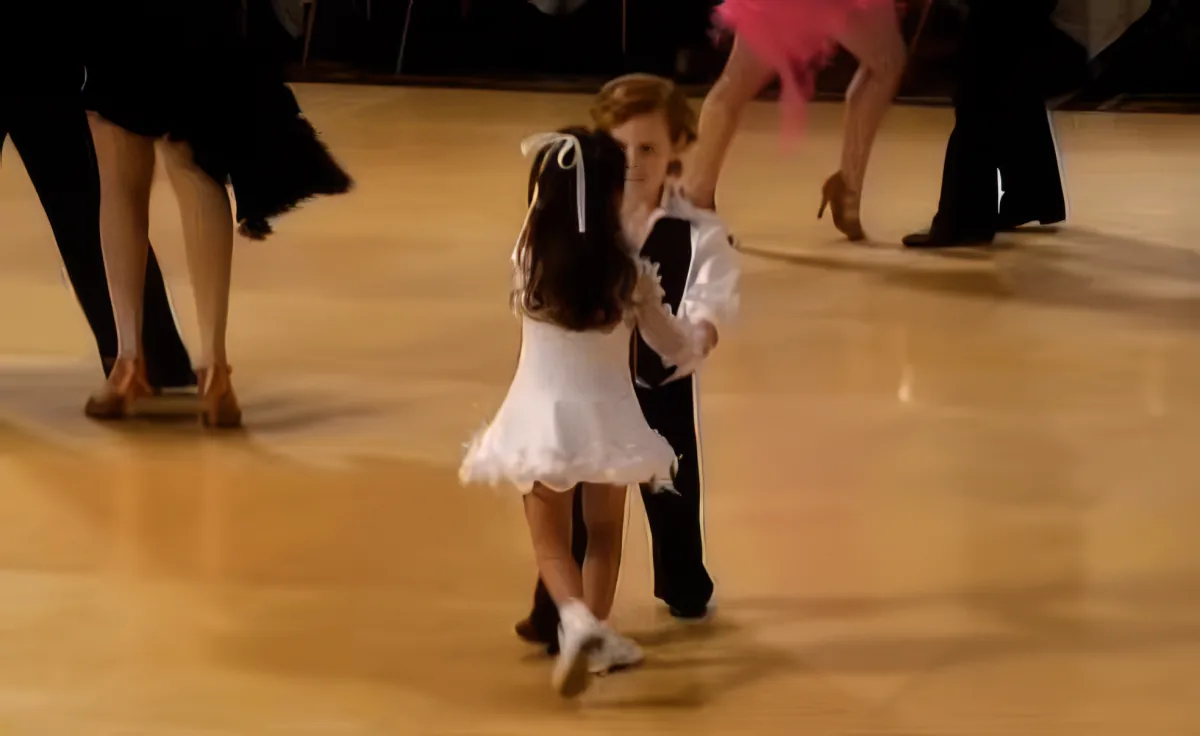 A young girl in a white dress and black skirt is dancing on a wooden floor, with another person in a white shirt and...