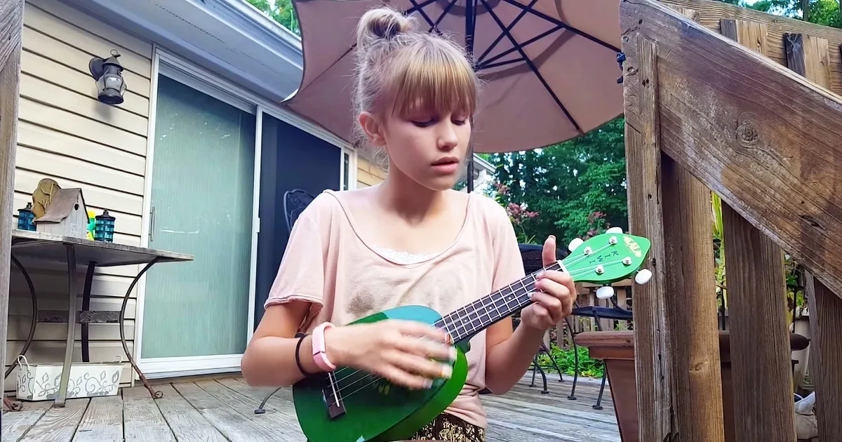 A young girl plays a green ukulele on a wooden deck under an umbrella.