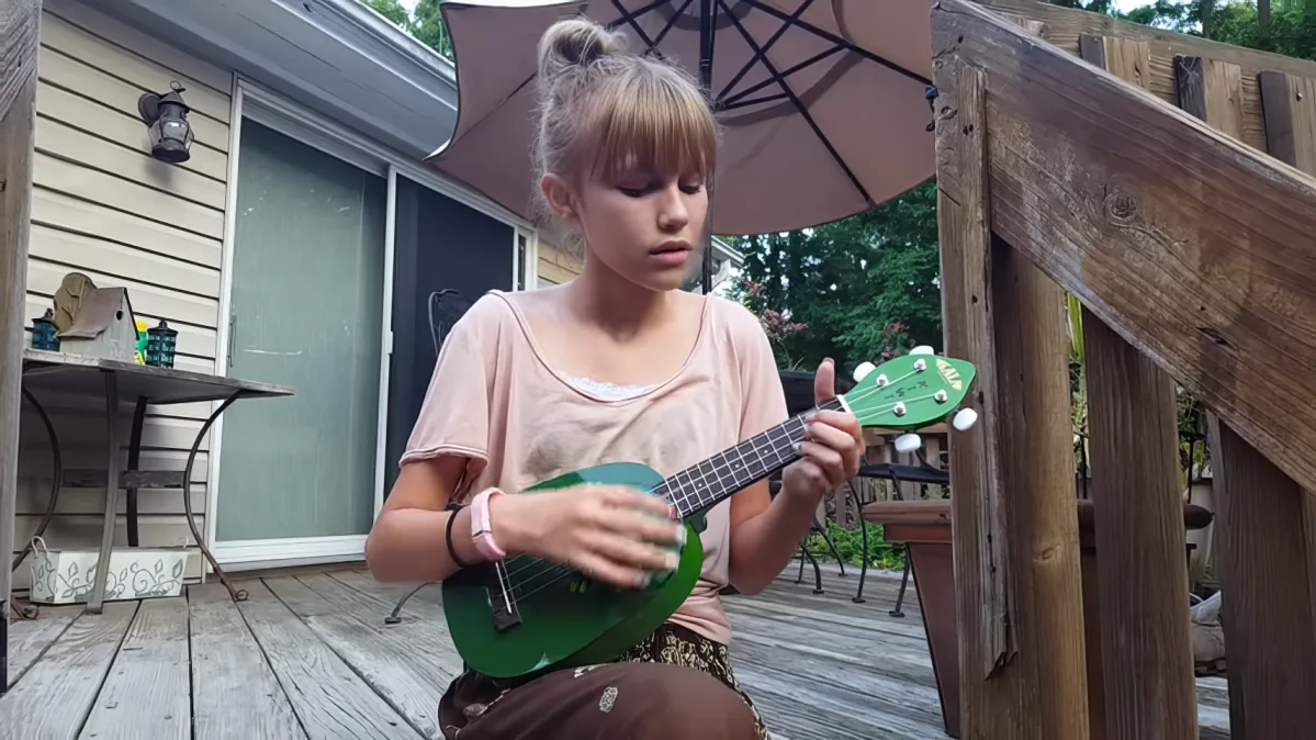 A young girl sits on a wooden deck, playing a green ukulele under a patio umbrella.