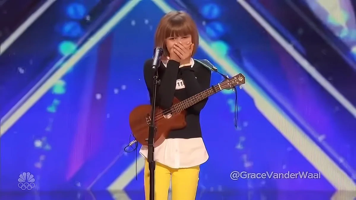 A young girl in yellow pants and a black top holds a ukulele, covering her mouth with her hand on stage.