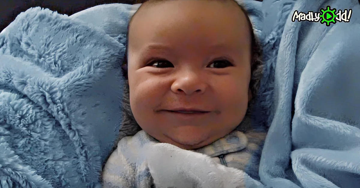 A smiling baby wrapped in a blue blanket, wearing a white outfit.