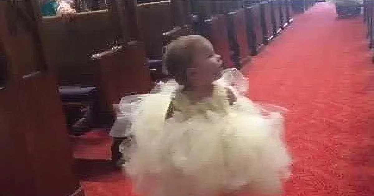 A baby in a white dress sits on a red carpeted church floor, surrounded by wooden pews.