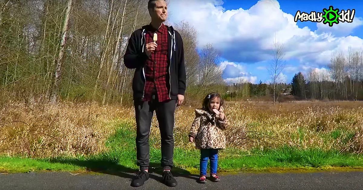 A man and a child stand on a road, both holding ice cream, with a grassy field and trees in the background.