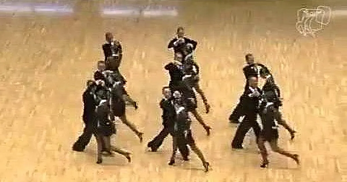 A group of dancers in formal attire performs a synchronized routine on a wooden floor.