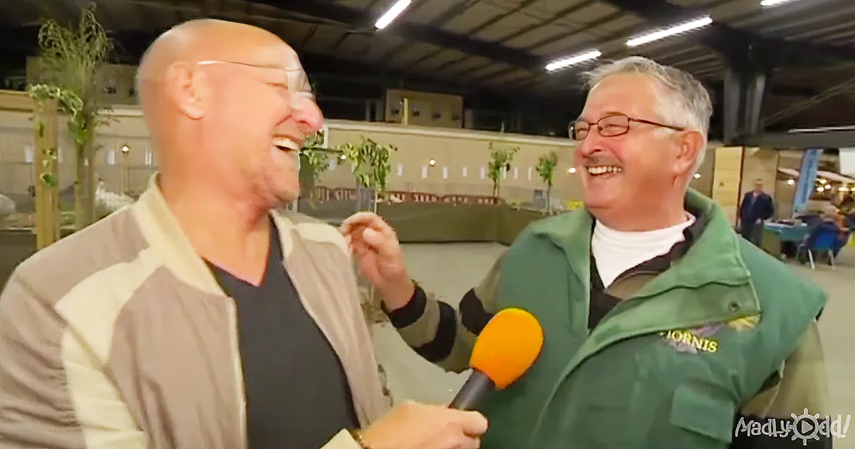 A man in a green jacket is interviewing another man in a beige jacket, both laughing.