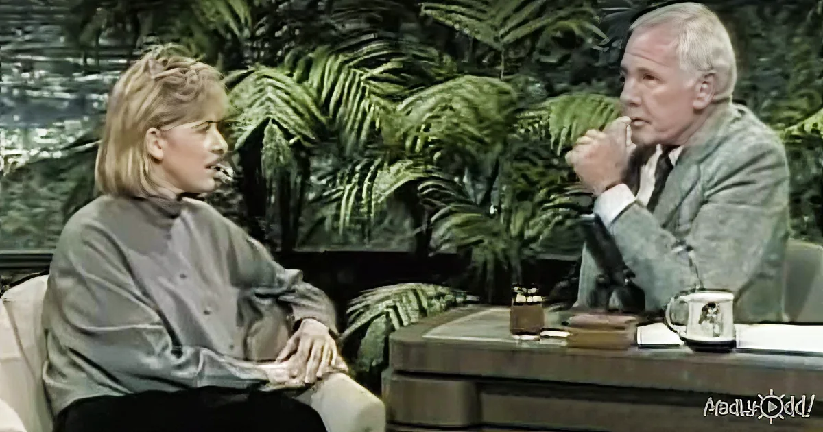 A woman in a gray turtleneck is seated, engaged in conversation with a man in a suit, both in a studio setting with...