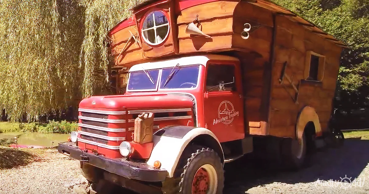 A vintage red truck with a wooden cabin on top is parked near a pond with trees in the background.