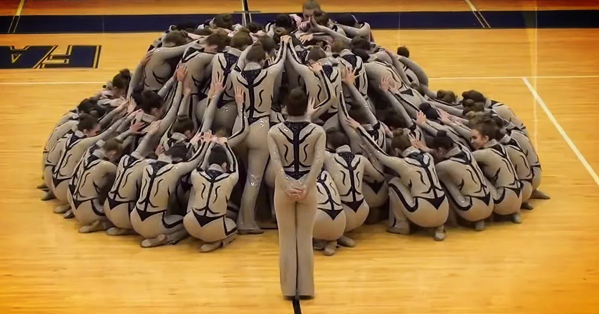A gymnast in a white leotard stands in front of a group of synchronized gymnasts in matching outfits, forming a pyramid...