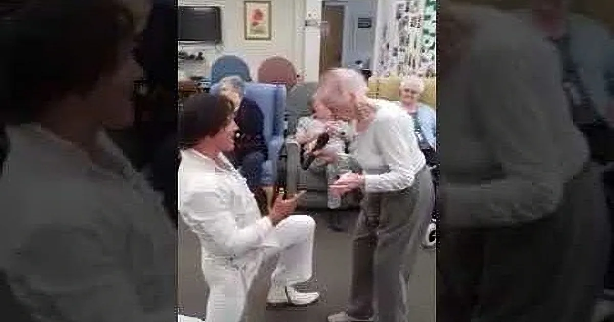 An elderly man in a white robe holds a baby, surrounded by people in a cozy indoor setting.