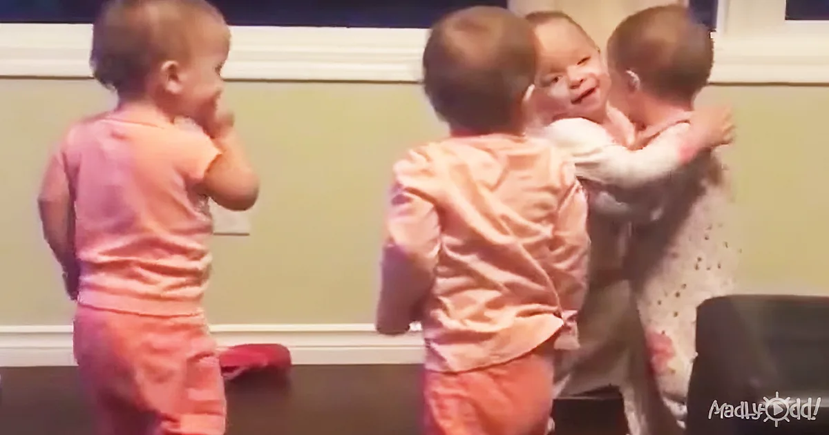 Two babies hug while another baby stands nearby, all wearing pink, in a room with a green wall.