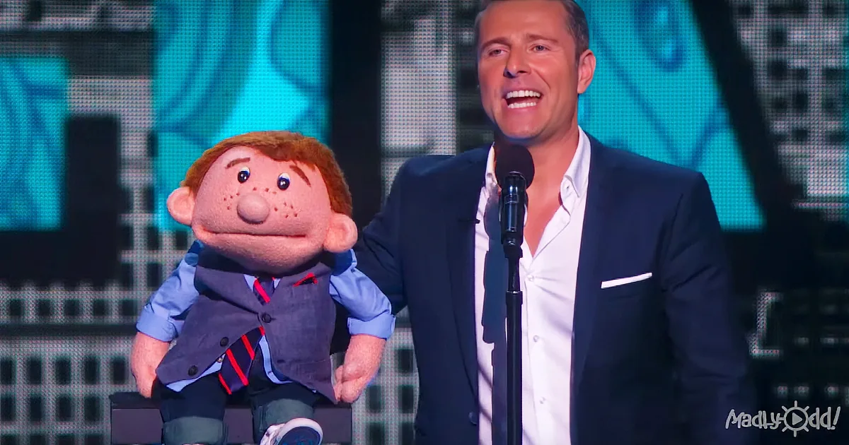 A man in a suit stands beside a puppet on stage, both smiling.