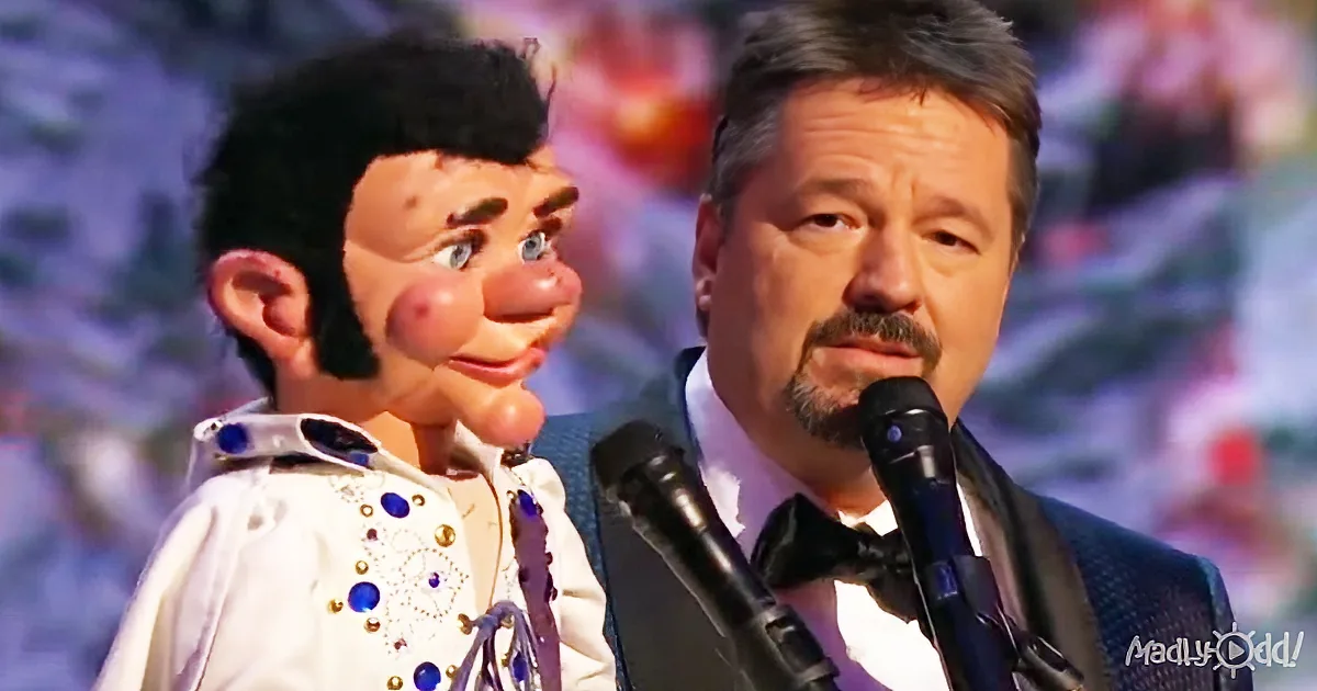 A man in a suit and bow tie stands beside a puppet dressed as Elvis Presley, both near microphones.