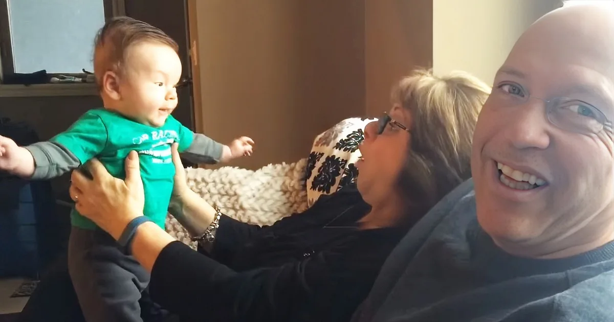 A baby in a green shirt is being held by a person in a black shirt, with another person smiling in the foreground.