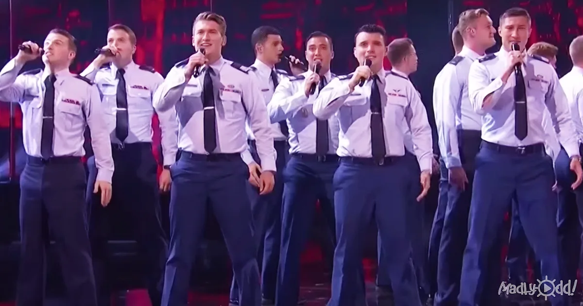A group of men in uniform sing on stage, holding microphones, with a red background.