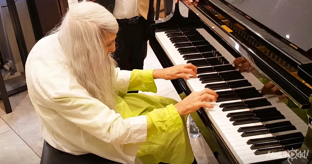 An elderly man with long white hair and beard plays a grand piano, wearing a white and green outfit.