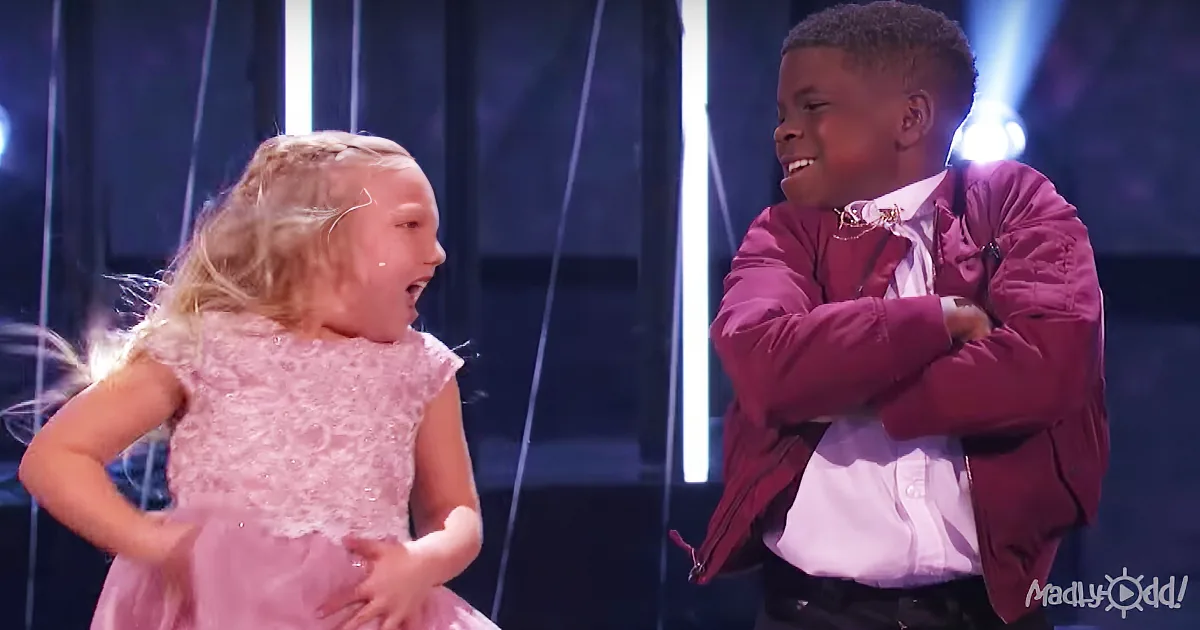 A young girl in a pink dress dances while a boy in a maroon jacket watches with a smile.
