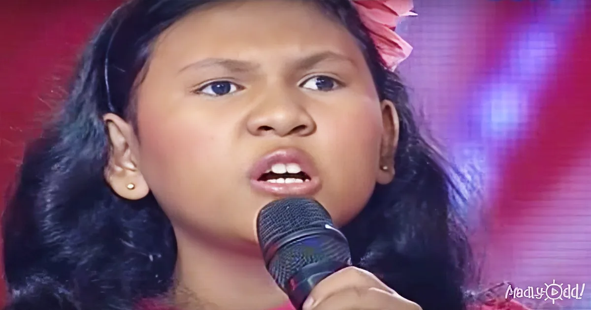A young girl with dark hair and earrings sings into a microphone, her mouth open mid-song.