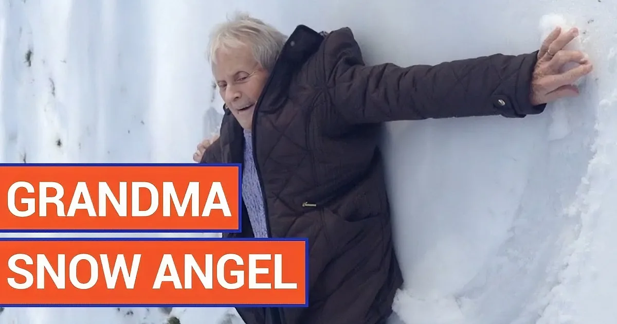 An elderly woman in a brown jacket is making a snow angel, smiling and enjoying the snow.