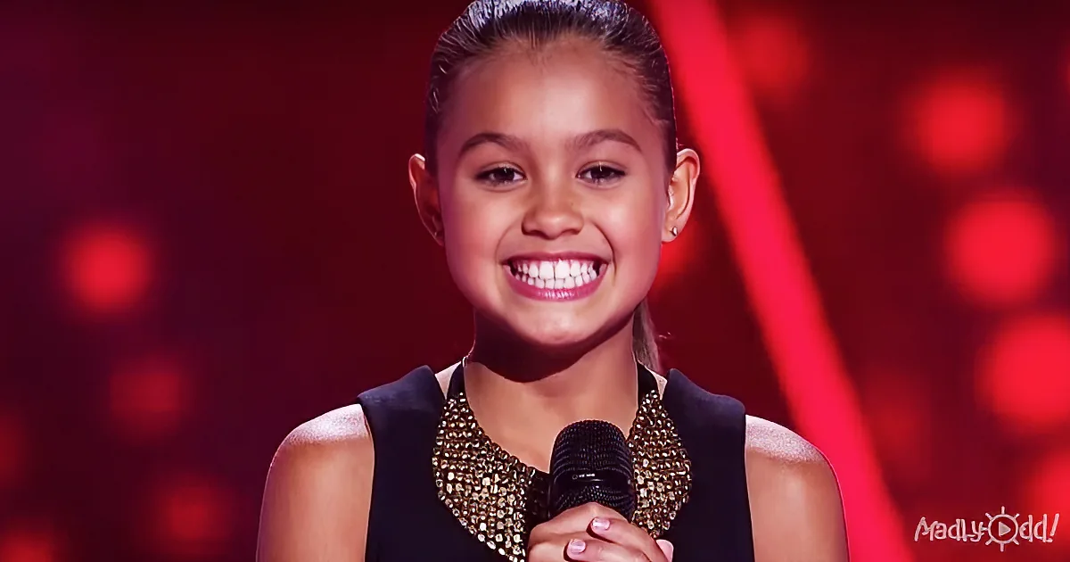 A smiling girl in a black dress with a gold necklace holds a microphone on a stage with red lights.