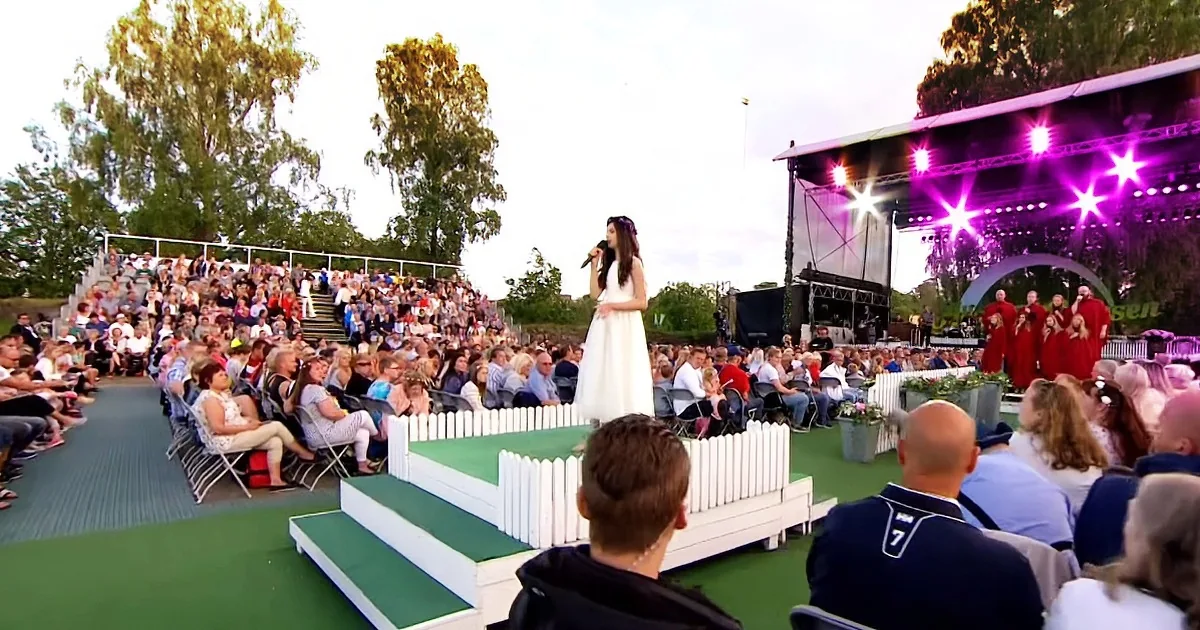 A woman in a white dress sings on a stage surrounded by a large audience under purple stage lights.