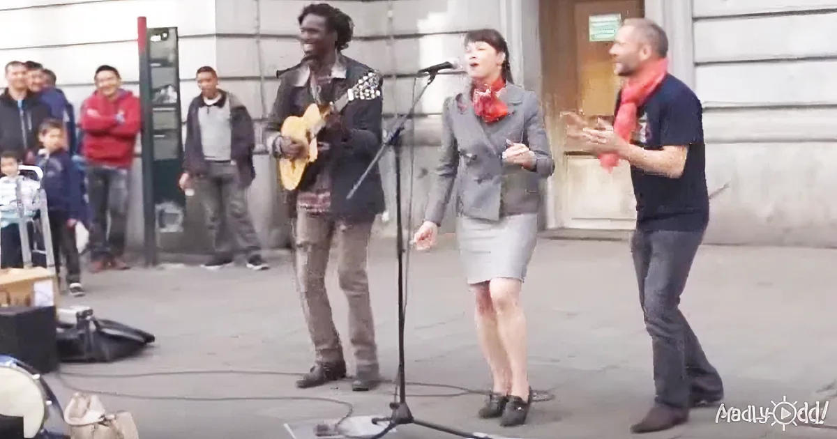 A woman sings into a microphone while a man plays guitar and another man gestures beside her.