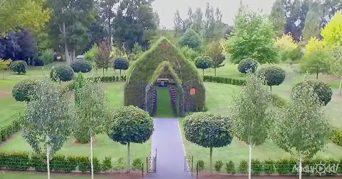 A whimsical, grass-covered structure resembling a house stands in a lush garden with neatly trimmed hedges and trees.