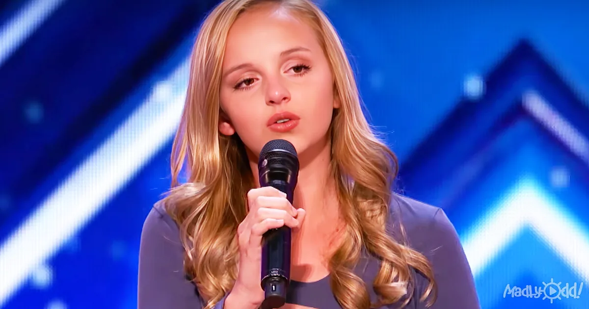 A woman with long blonde hair holds a microphone, standing in front of a blue background with geometric shapes.