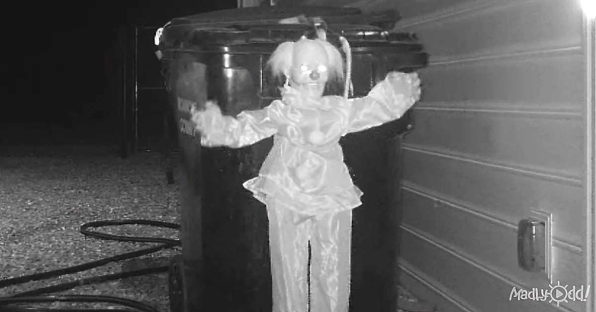 A creepy doll with outstretched arms stands in front of a trash can and garage door at night.