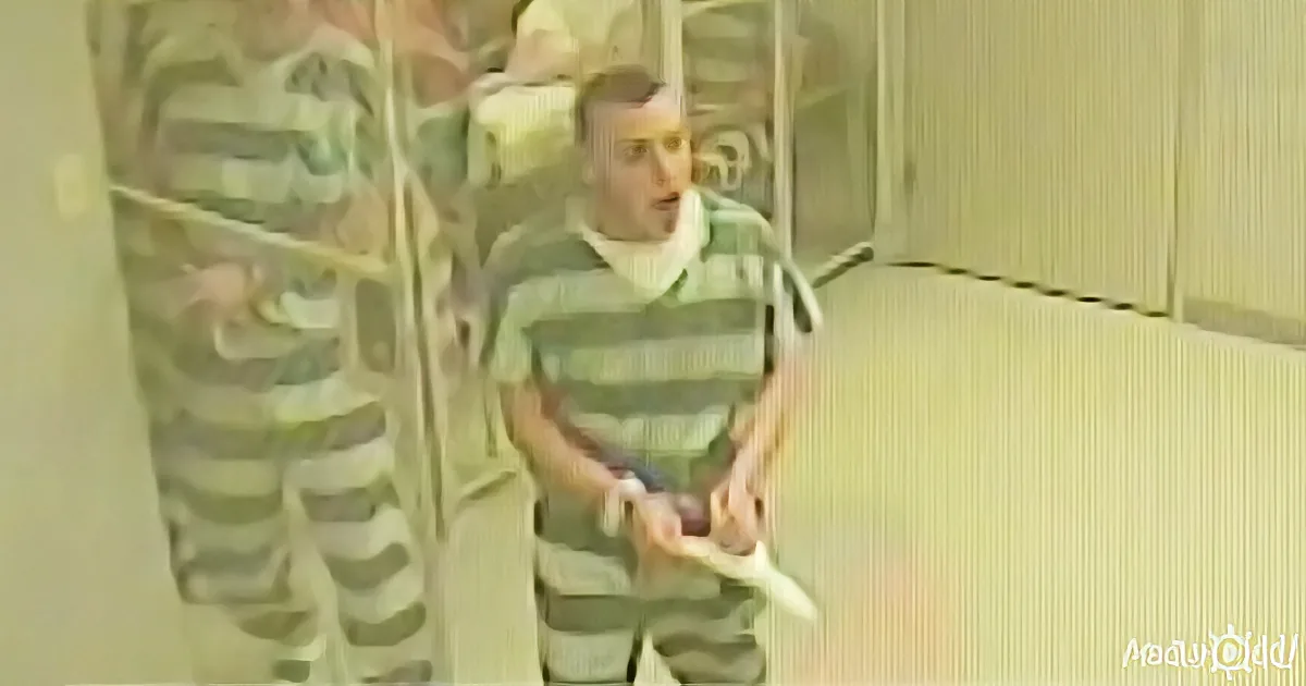 A man in a striped uniform stands in a room, holding a small object, with another person partially visible behind him.