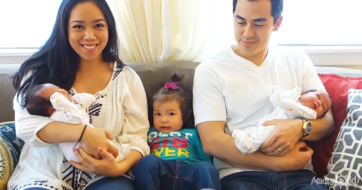A family of four sits on a couch, with two adults holding newborn babies and a toddler in the middle.