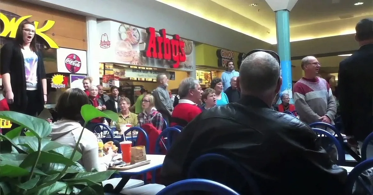 A woman sings in a crowded mall food court with Arby's and other restaurants visible.