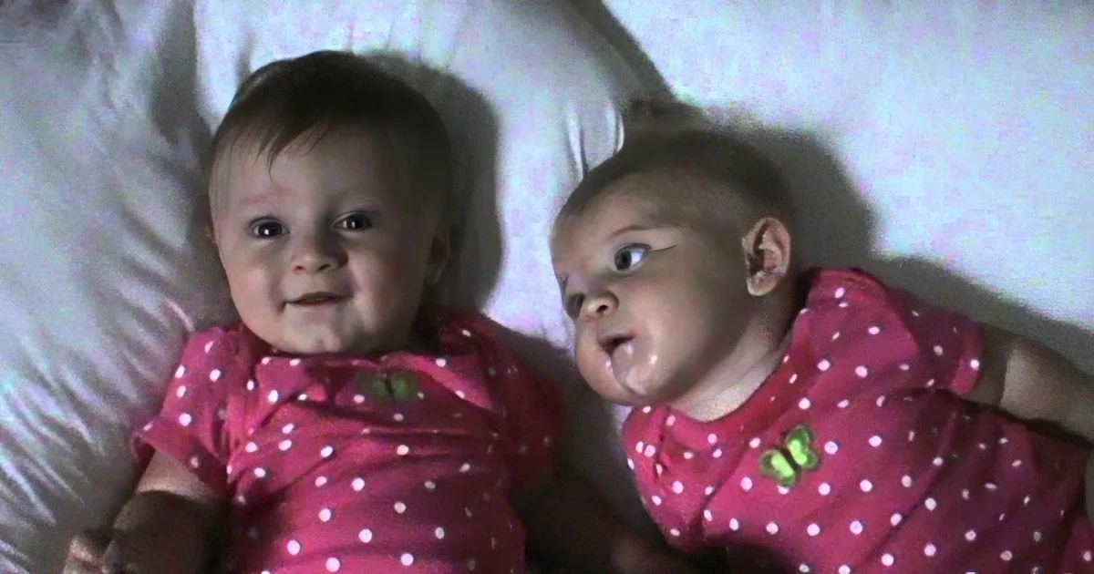 Two babies in matching pink polka-dot pajamas are lying on a white bed, smiling and looking at the camera.