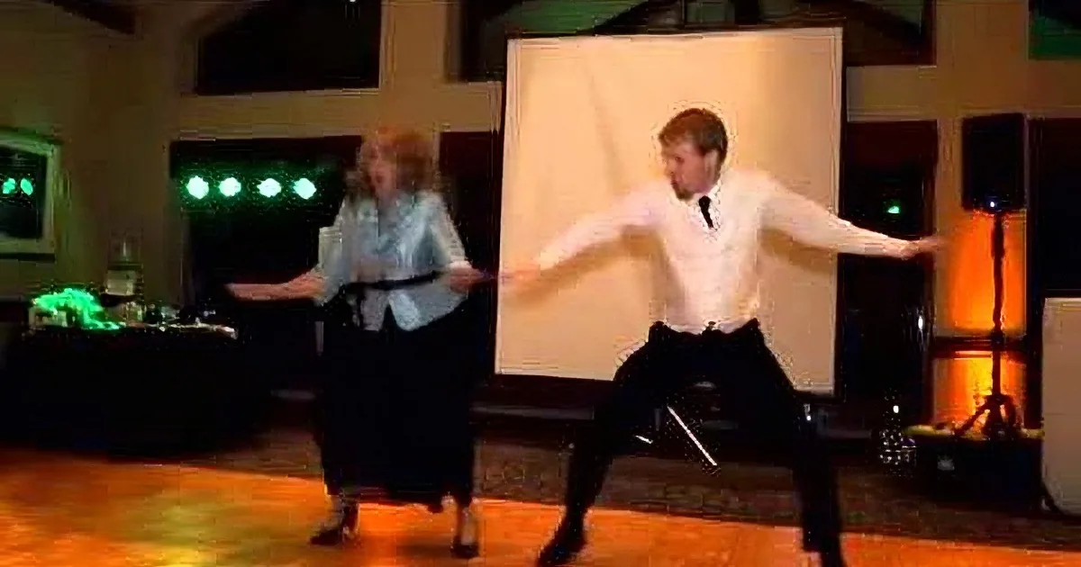 A man and woman are dancing energetically on a wooden floor, with a backdrop of stage lights and a white curtain.