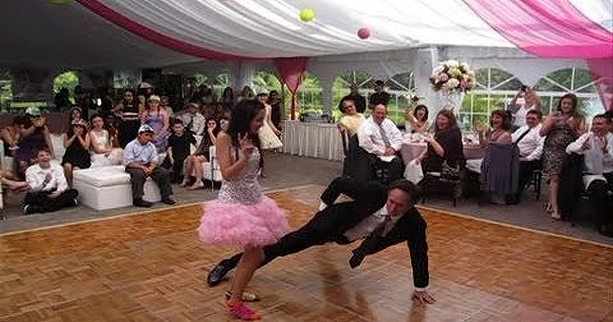 A woman in a pink tutu dances energetically on a wooden floor, while a man in a suit supports her with a dramatic pose.