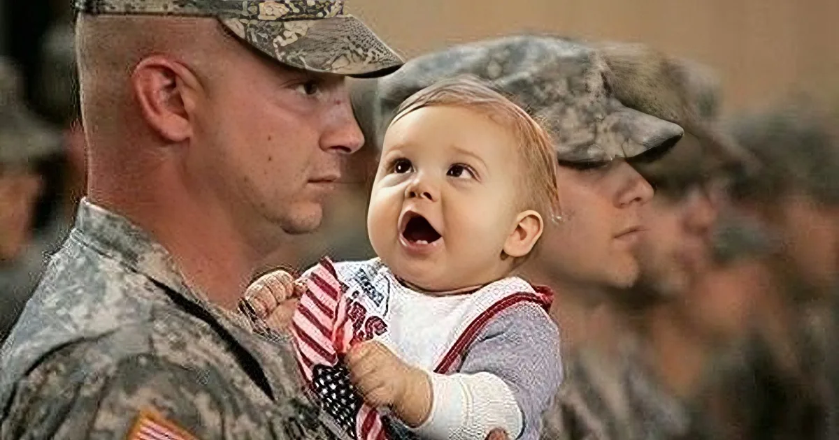 A soldier in uniform holds a baby, both looking at each other, with other soldiers in the background.