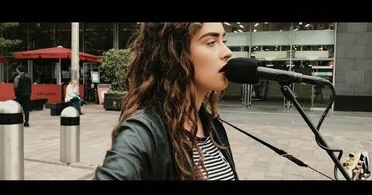 A woman with long, curly hair sings into a microphone in an urban setting with people and buildings in the background.