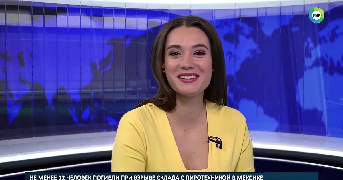 A woman in a yellow dress smiles on a news set with a world map background.