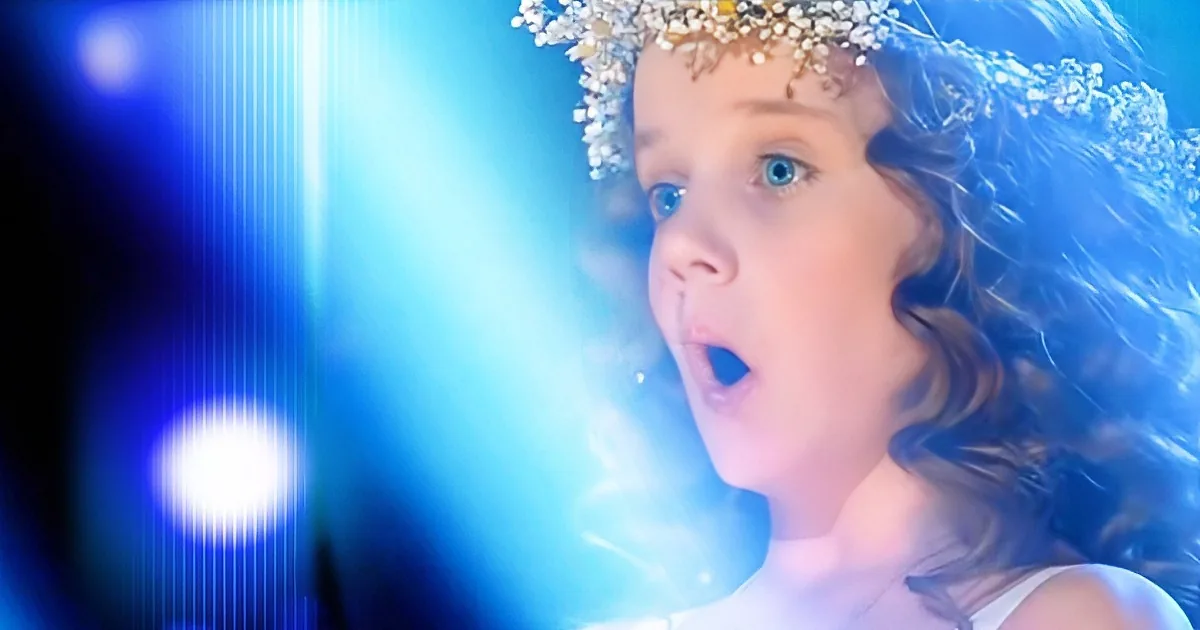 A young child with curly hair and a floral crown, mouth open, against a bright blue background.