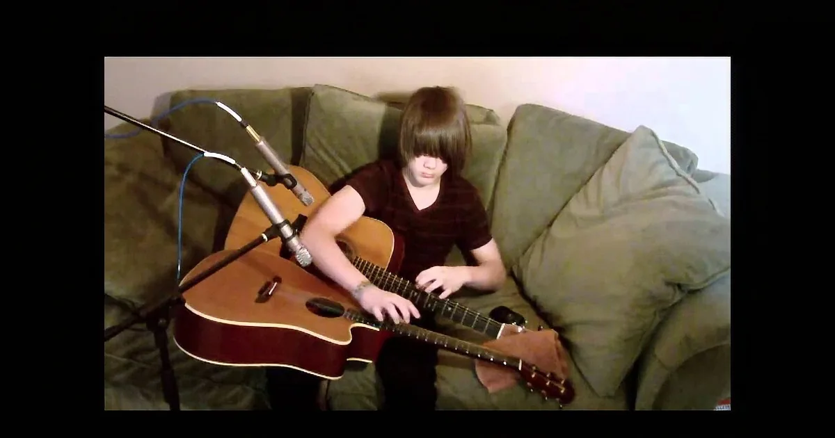 A person sits on a green couch playing an acoustic guitar with a microphone in front.