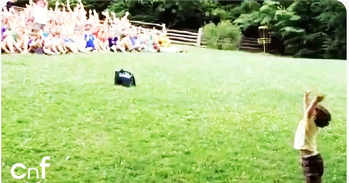 A young boy raises his arms in a grassy field, with a crowd of people watching in the background.